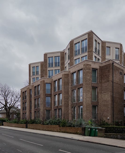 A modern multi-storey residential building with a combination of brick and light-colored facades, featuring large rectangular windows and small balconies. The building is situated along a paved street